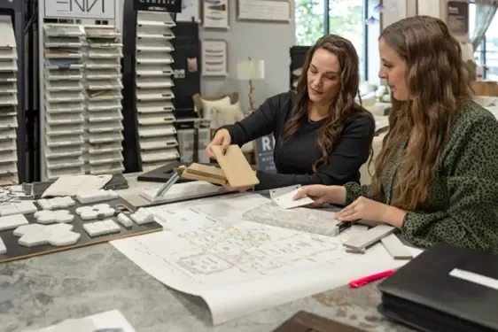 Two women review architectural or design plans at a table, with one pointing to material samples displayed on shelving behind them