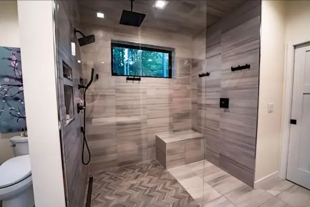 Modern bathroom shower enclosure with gray tile walls, glass door, built-in bench, rainfall and handheld showerheads, and window overlooking trees